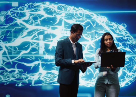 Un uomo e una donna in piedi che guardano lo schermo del pc, sullo sfondo l'immagine digitale di un cervello