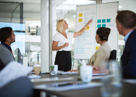 Giovane donna presenta ai colleghi, sullo sfondo una lavagna con dei post it