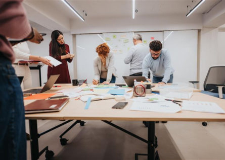 Gruppo di persone lavora in team sopra grande tavolo in legno, sullo sfondo una lavagna con dei post it