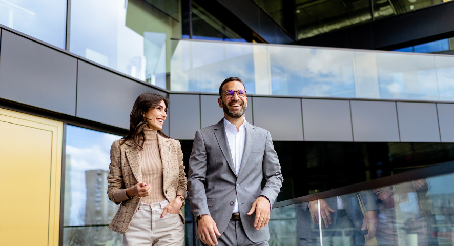 Una professionista e un professionista caminnano sorridendo, sullo sfondo vetri come di grattacieli